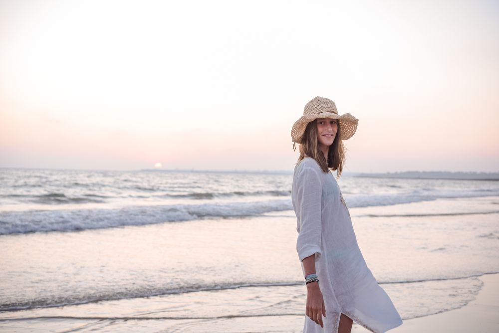 Charming woman in light white dress looking at camera over shoulder on wavy beach