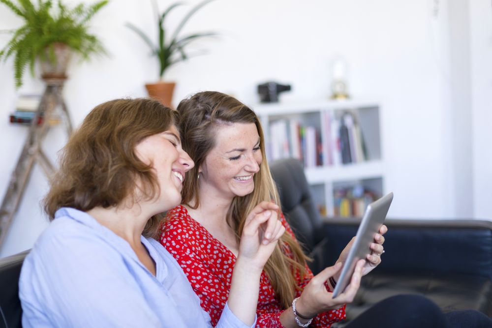 Two laughing women at home sitting on sofa using digital tablet