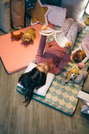 Happy little girl reading and relaxing lying over sleeping mat in tent surrounded by stuffed dolls