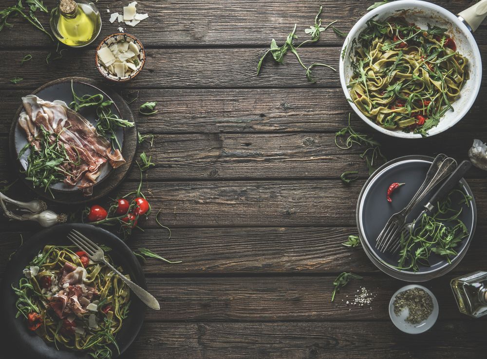 Green pasta on plates, ingredients and kitchen utensils with Parma ham, tomatoes, arugula, cheese and olive oil