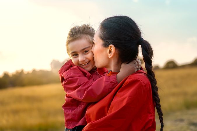 Mother affectionately kissing her happy daughter outdoors at sunset.