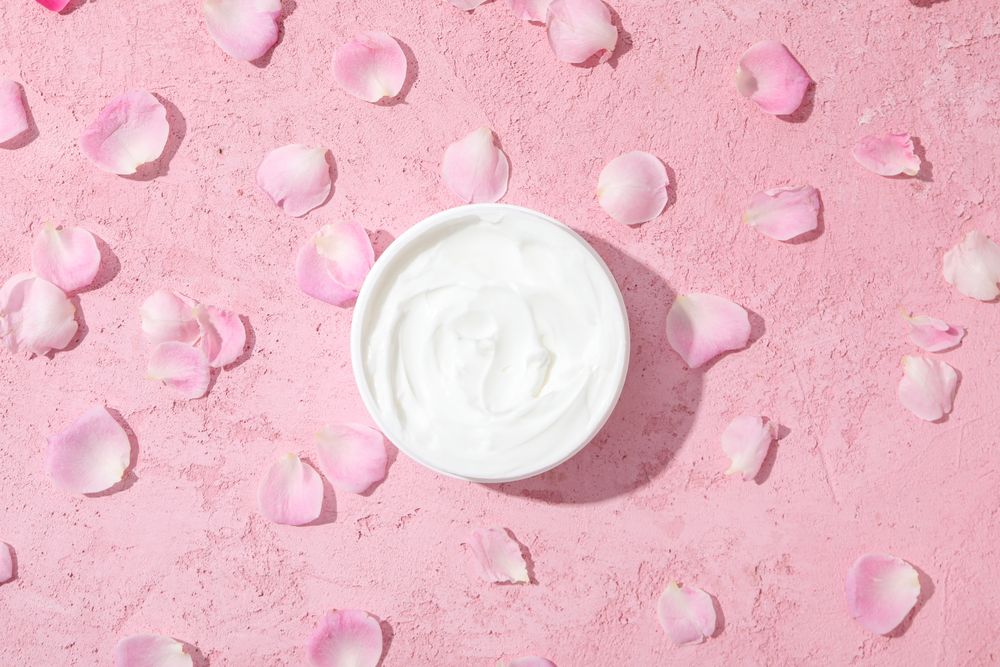 Jar with cream and rose petals on pink background, top view