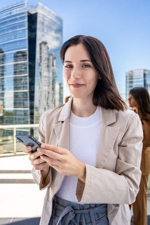 A woman dressed in business attire is standing outside with a cell phone in her hand and about to use it