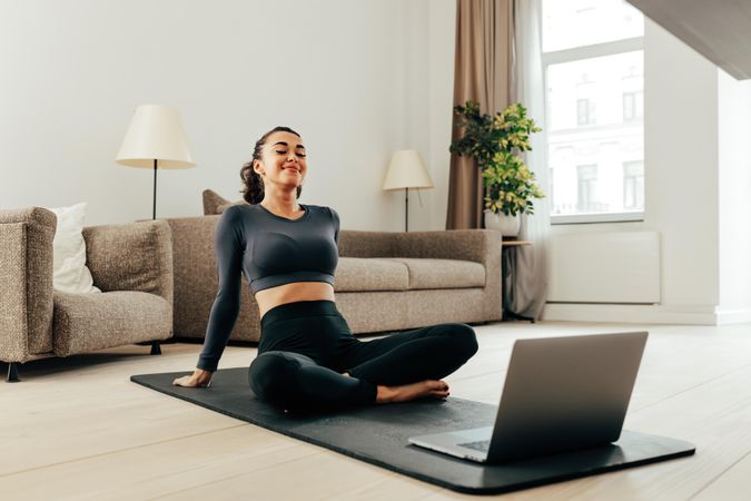 Woman watching exercise video on her laptop sitting on a yoga mat