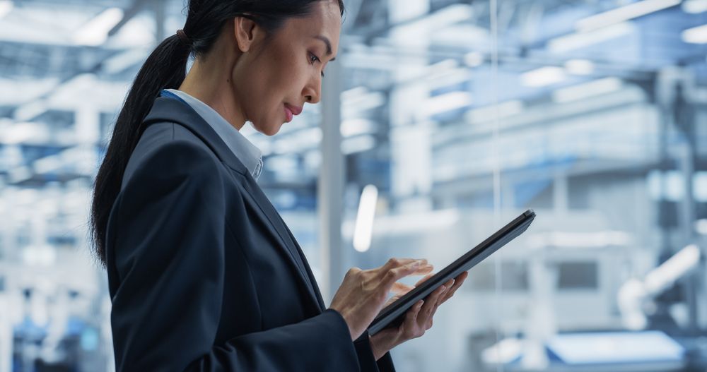 Asian Female Engineer Standing Next to a Window in Office, Using Tablet Computer at an Electronics Manufacture. Specialist Using an Online Software to Program Robots on Assembly Line