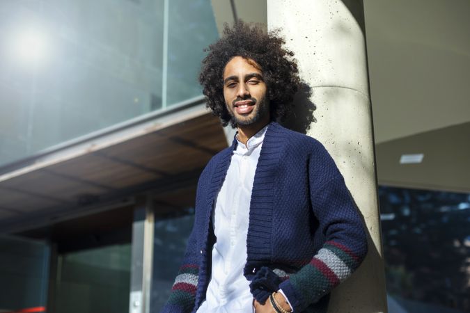 Portrait of Black man in cardigan and jeans leaning on pillar outside on sunny day