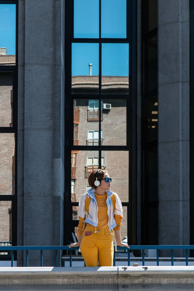 Stylish pensive young woman in sunglasses and headphones standing on road bridge in city