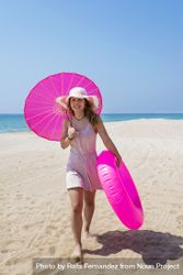 Smiling woman in pink dress walking on sand beach wearing a hat and holding a parasol and a floater 4d89Ll