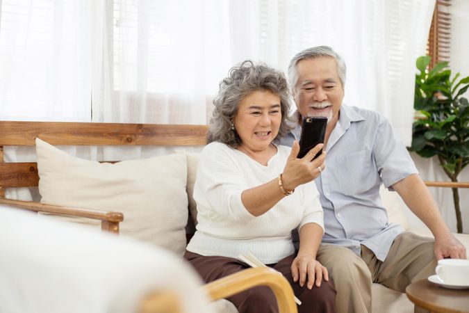 Happy Asian couple sitting on sofa using smartphone video call chatting with family