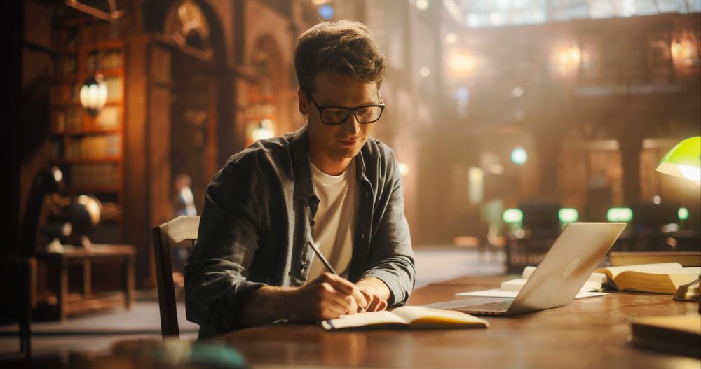 Concentrated Young Man Writing in Notebook at a Vintage Library Desk, With Laptop Open for Online Research