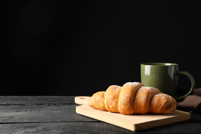 Side view of board with croissant and mug of coffee on dark wooden background, copy space