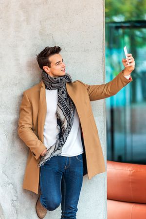Stylish man leaning on wall outside taking selfie in camel coat 