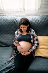 A young woman with a very advanced pregnancy is sitting on the couch looking at and talking on her cell phone.