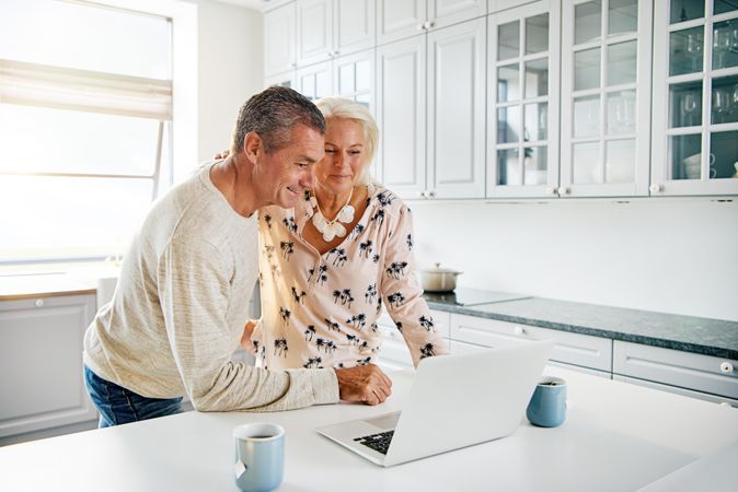 Older couple in bright kitchen with cups of tea and a laptop