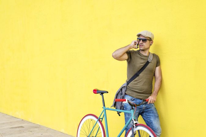 Male in hat and sunglasses standing next to yellow wall with bike and speaking on phone, copy space