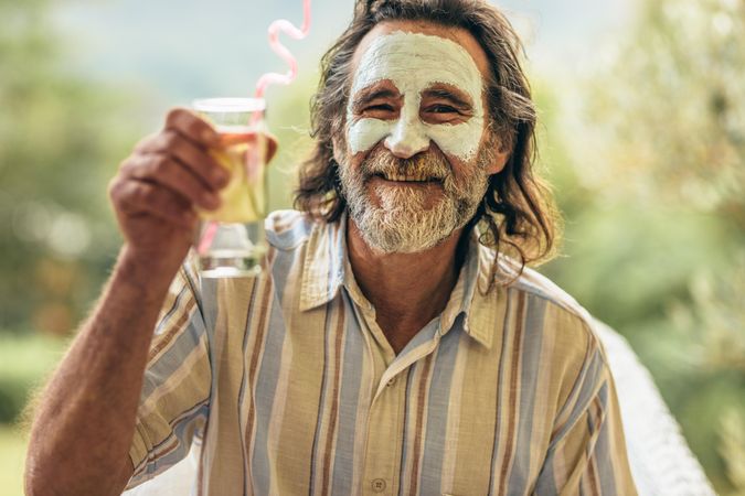 Bearded man with clay facial mask holding a glass of juice in hand