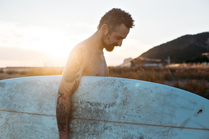 Tattooed man looking down with surfboard under his arm at sunset