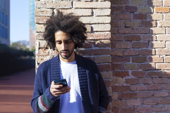 Curly haired man looking at his smartphone while leaning on a brick wall outdoors on sunny day