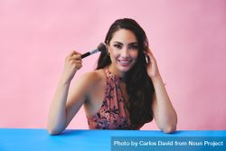 Elegant Hispanic woman with long brown hair holding large make up brush up to her face 561El0