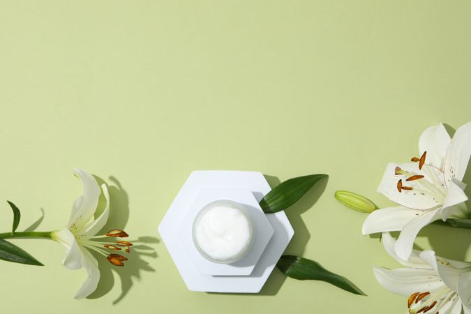 Jar with cream and lilies on green background, space for text