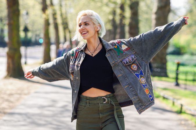 Blonde woman in jean jacket with arms outstretched while walking in park