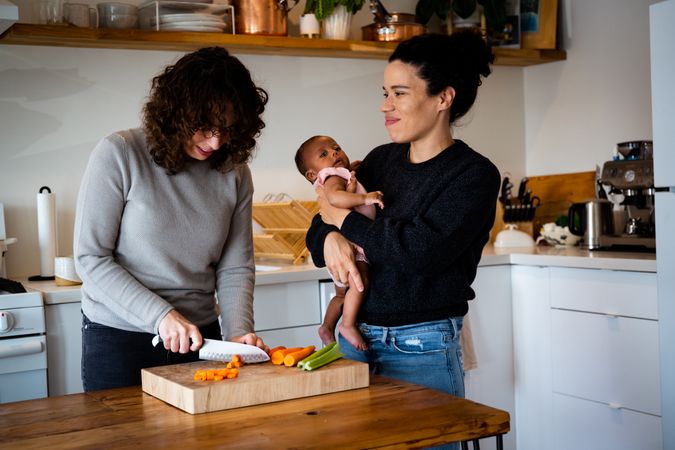 Two partners in the kitchen chopping vegetables with other holding baby