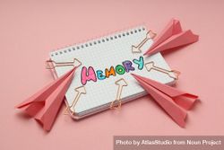 Notepad with “memory” written in colorful markers with arrow shaped paper planes and paper clips 5q711b