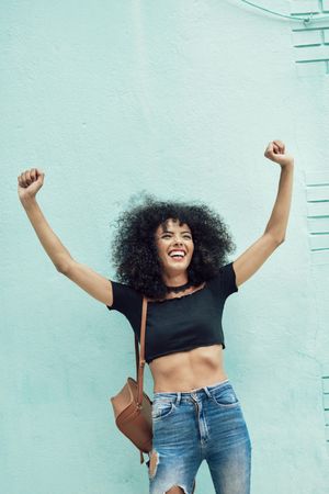 Female in casual clothes leaning on turquoise wall and smiling while raising her arms in celebration