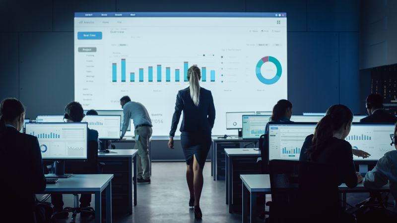 Data Analysts Work in Dimly Lit Room With Large Screen Displaying Financial Charts.
