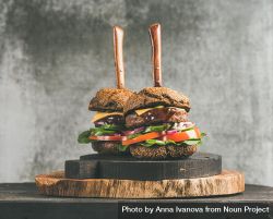 Two cheeseburgers skewered with knives, with fresh vegetables, on wooden board 4Ajwqb