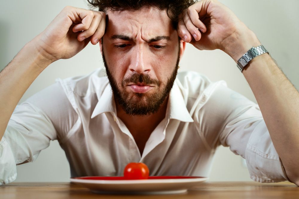 Frustrated man on a strict diet looking at a single cherry tomato on a plate.