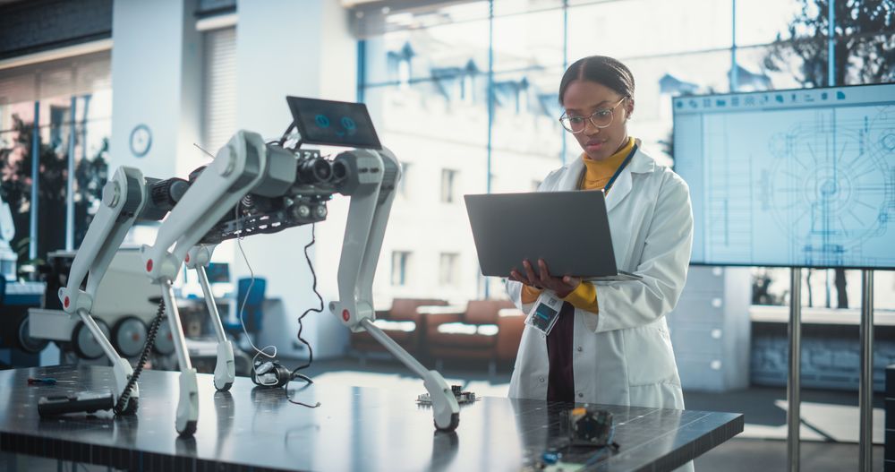 Portrait of Young Black Female Specialist in Lab Coat Using Laptop to Test an AI Robotic Prototype.