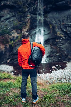Back view of a man in red jacket with backpack standing facing waterfall