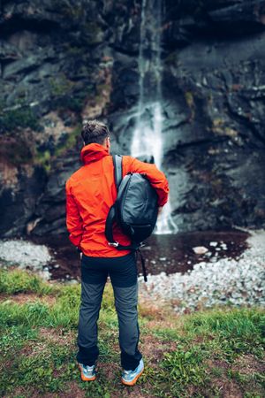 Back view of a man in red jacket with backpack standing facing waterfall
