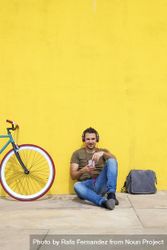 Male sitting in front of yellow wall next to bike and listening to music on smartphone, vertical 0WqzP4