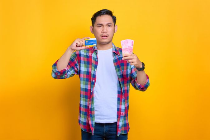 Upset Asian man holding credit card and cash in studio shoot
