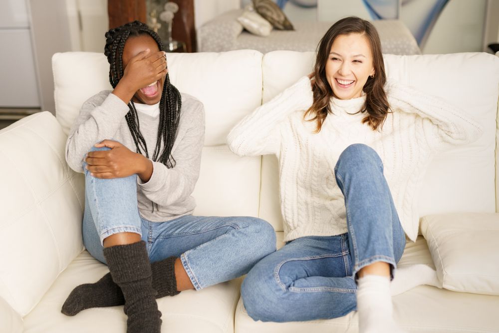Two women in jeans and sweater joking together on a sofa