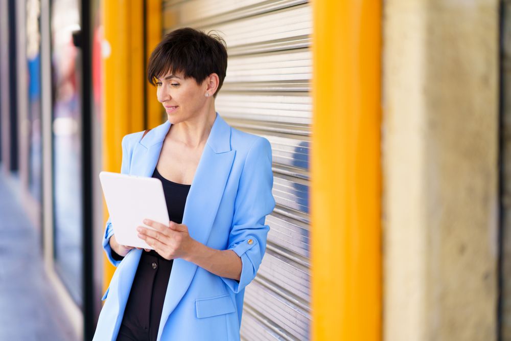 Female in blue blazer leaning on shutter outside reading digital tablet