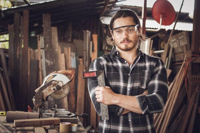 Portrait of male woodworker wearing safety glasses and holding tool in workshop surrounded by wood