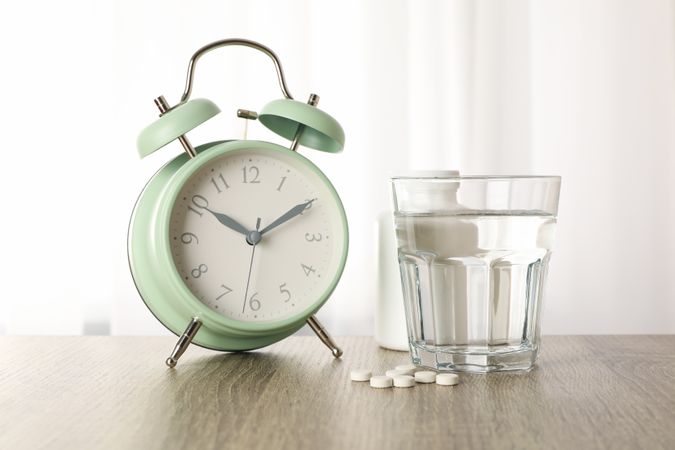 Glass of water with pills next to alarm clock next to window in beige room