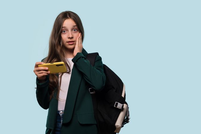 Surprised teen girl holding phone, wearing backpack, thoughtful expression.