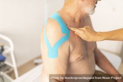 A female physiotherapist checks the tape from the shoulder of her patient, an older man 5Q226g