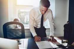 Man in shirt and tie standing over desk and writing in sunny office