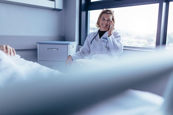 Happy female doctor sitting in hospital room and making a phone call