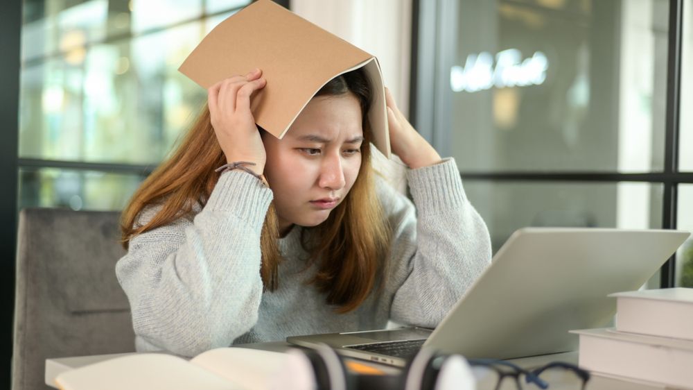 Asian woman holding a book on her head stressed looking at laptop during online lessons from laptop.