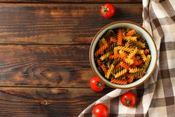 Bowl with color pasta, tomatoes and kitchen towel on wooden background, space for text