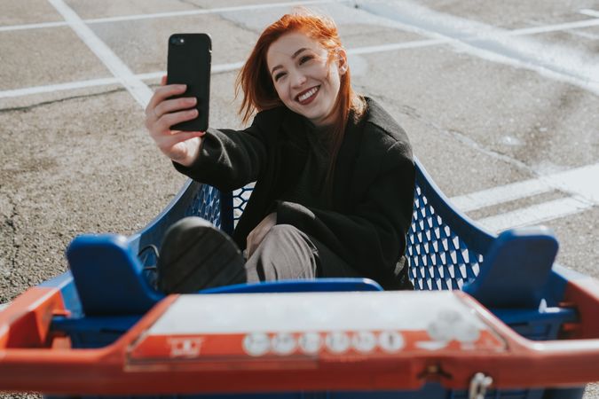 Smiling woman with smartphone in shopping trolley in parking lot