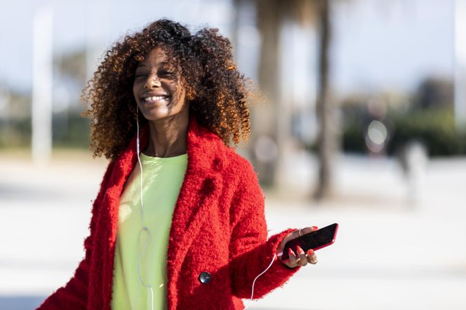 Smiling female in red jacket listening to music on phone standing on street with palm trees