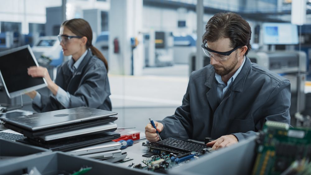 Male And Female Workers Taking Apart Old Laptops To Recycle Electronic Components For Microchip Production At Modern Electronics Factory.
