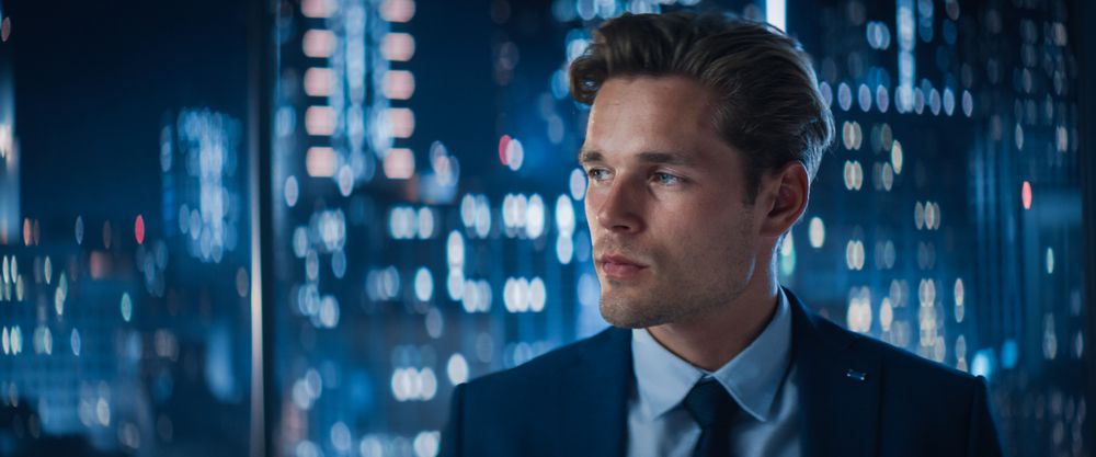 Close Up Portrait of Entrepreneur in Stylish Suit Posing Next to Window in Big City Office with Skyscrapers Late At Night.